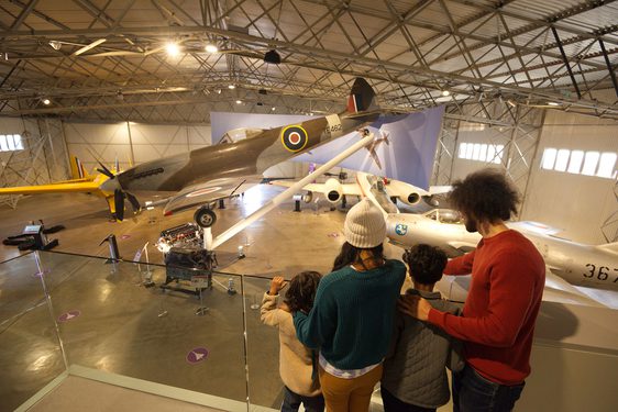 A family of four look down onto several military aircraft from a viewing platform within a hangar.