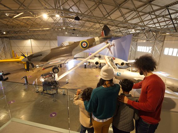 A family of four look down onto several military aircraft from a viewing platform within a hangar.