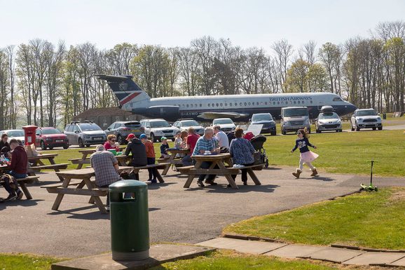 People sitting on picnic tables next to a car park. There is a passenger aircraft parked in the background.