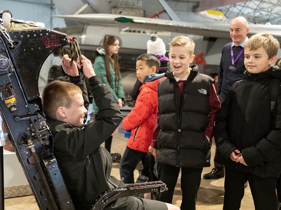 A school pupil sitting in a pilot seat with other pupils looking on.