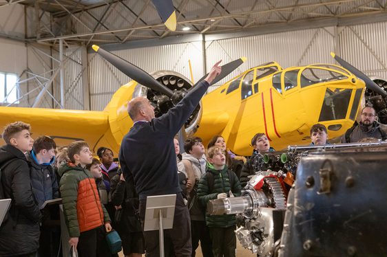 A group of school pupils in an aircraft hangar listening to a teacher talking.