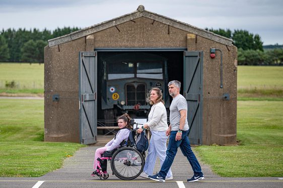 Two parents and a child in a wheelchair walk through a historic airfield. They walk by a vintage military truck in a storage shed.