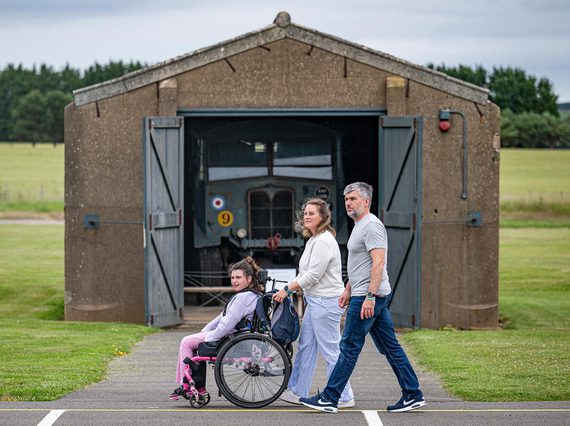 Two parents and a child in a wheelchair walk through a historic airfield. They walk by a vintage military truck in a storage shed.