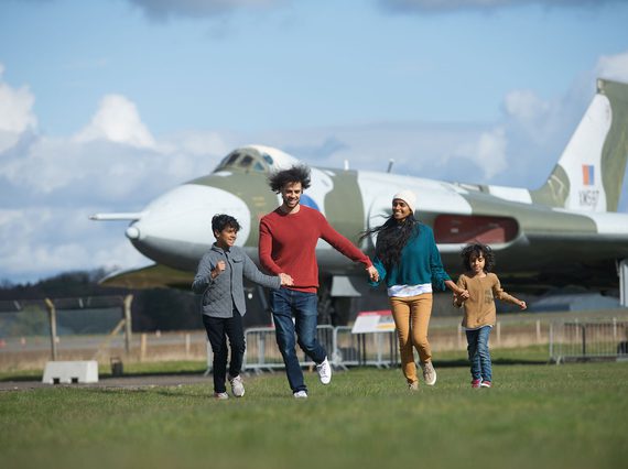 A family of four holding hands, running in a grassy field, with a large Vulcan aircraft in the background.