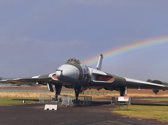 A Vulcan aircraft stationed in an airfield outside a black aviation hangar. There is a rainbow in the sky in the background.