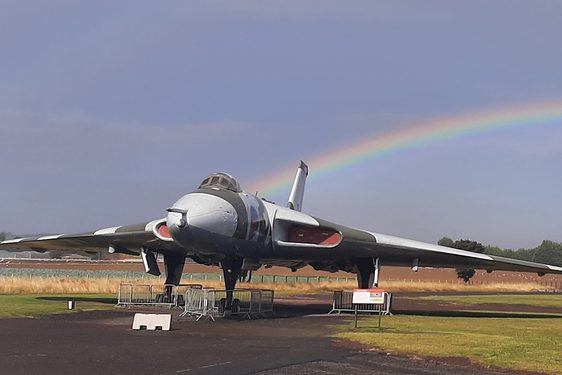 A Vulcan aircraft stationed in an airfield outside a black aviation hangar. There is a rainbow in the sky in the background.