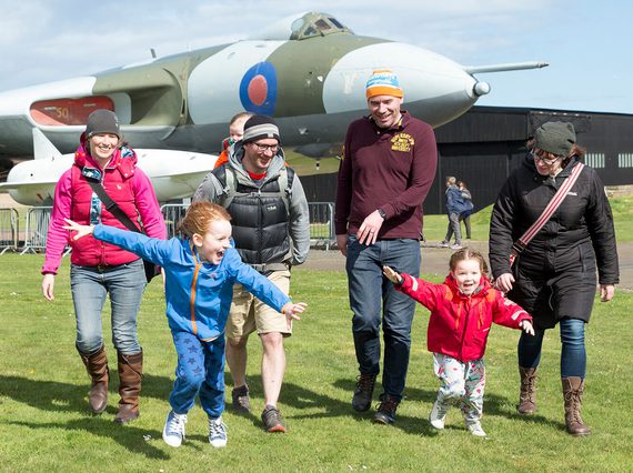 A family walking in an airfield away from a Vulcan aircraft. Two children are running around pretending to be aeroplanes.