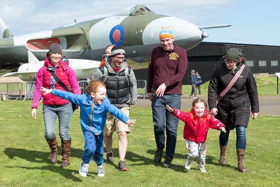 A family walking in an airfield away from a Vulcan aircraft. Two children are running around pretending to be aeroplanes.