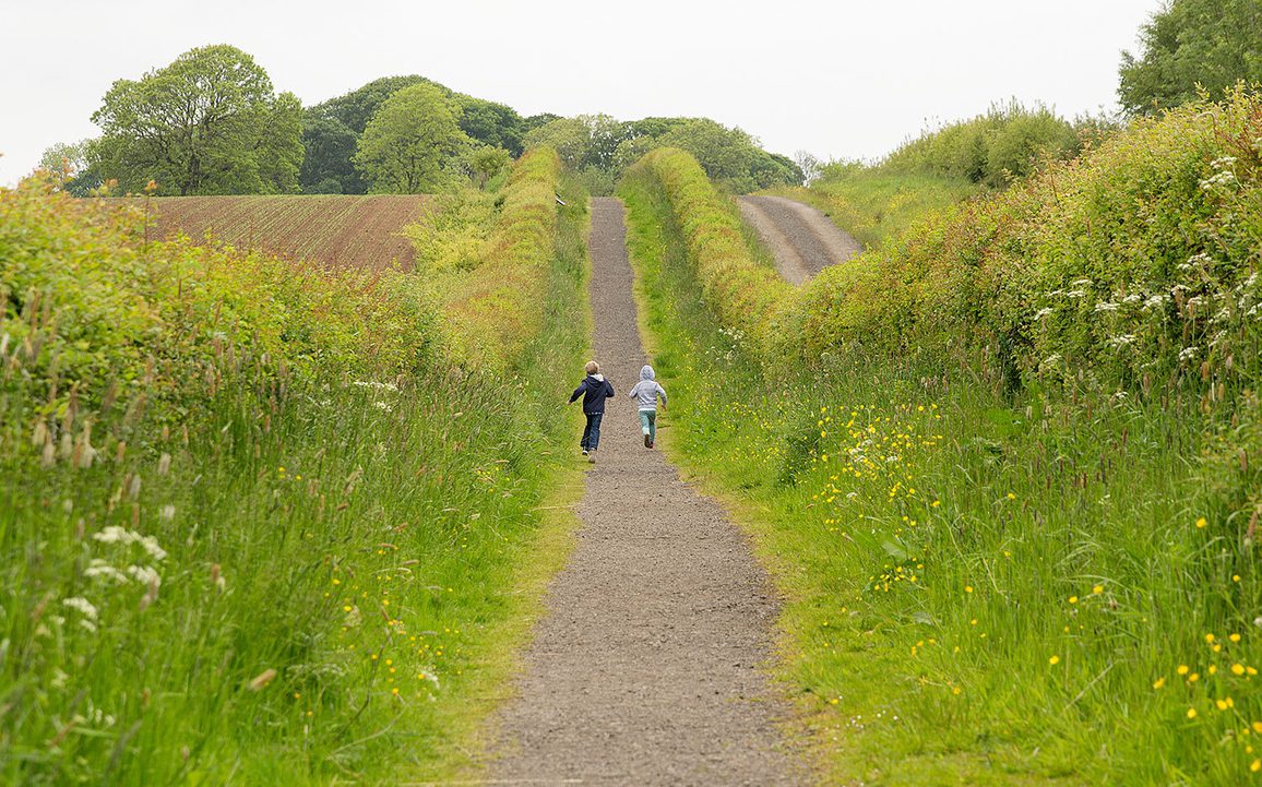 Two children run down a hedge-lined path ion the countryside.