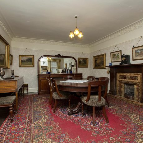 The interior of a vintage dining room, with ornate wooden furniture and framed portraits on the walls.