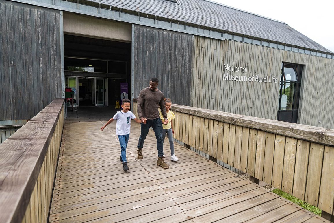 A family of three holding hands and walking along a footbridge away from the entrance to the National Museum of Rural Life.