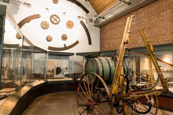 A museum gallery with tall brick walls. Museum cases are arranged in a semi-circle around a wooden carriage.