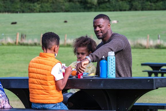 A family sitting at a bench in a playpark eating lunch