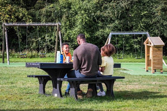 A family of three sitting at a bench in a playpark eating lunch.