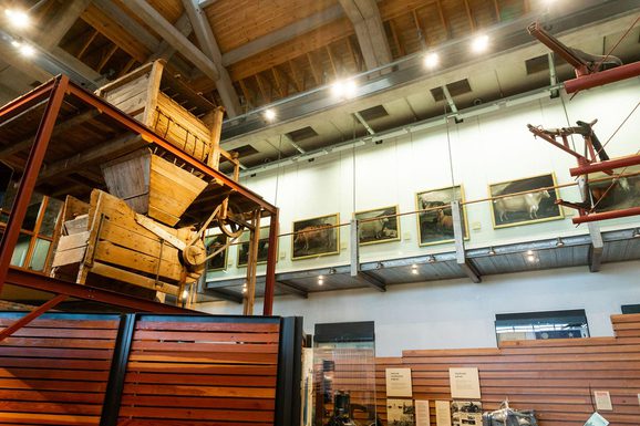 A large gallery with agricultural tools and portraits. A upper level walkway spans the width of the gallery where the portraits hang.