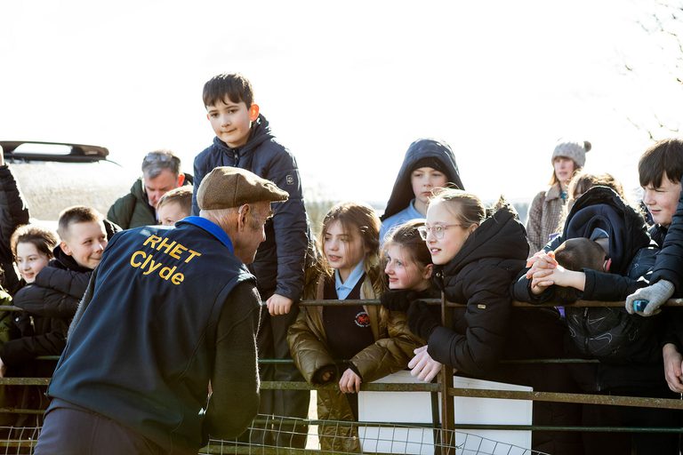 A group of school children on a farm lean over a fence to talk to a farmer.