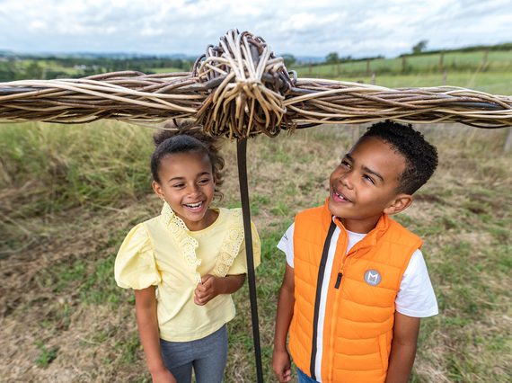 Two children standing underneath a bird sculpture made of willow.