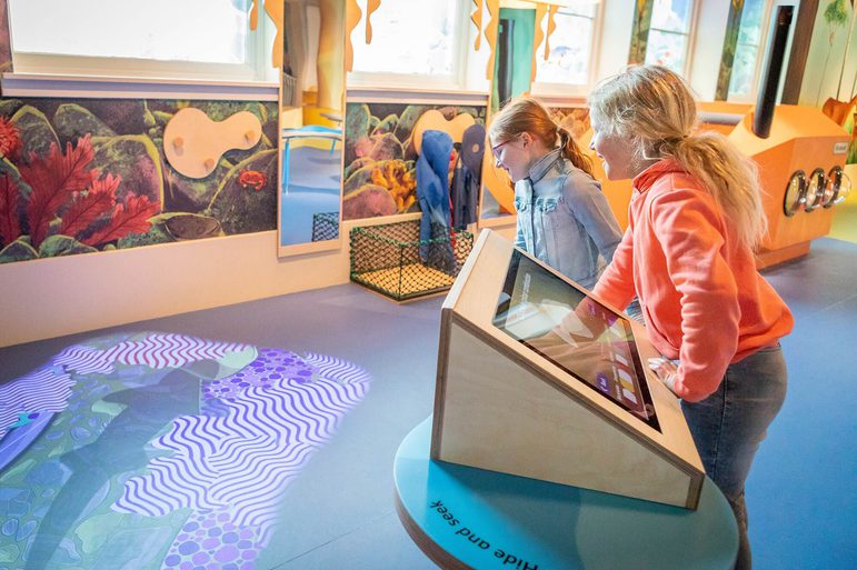 Two young children play with an interactive screen in a colourful, playful space. They are looking at a projection of cartoon fish on the floor.