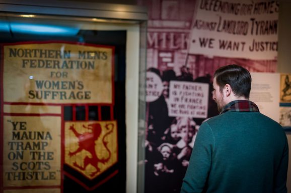 A museum visitor looking at a large flag that reads "Northern Men's Federation for Women's Suffrage."
