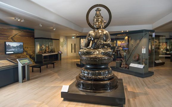 View of a museum gallery with a large bronze sculpture of a buddha in the center.
