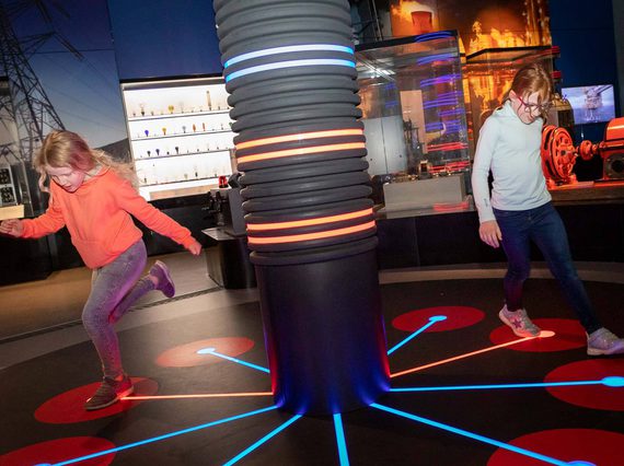 Two children run around a lighted pillar in a museum gallery, stepping on different coloured lights on the floor.