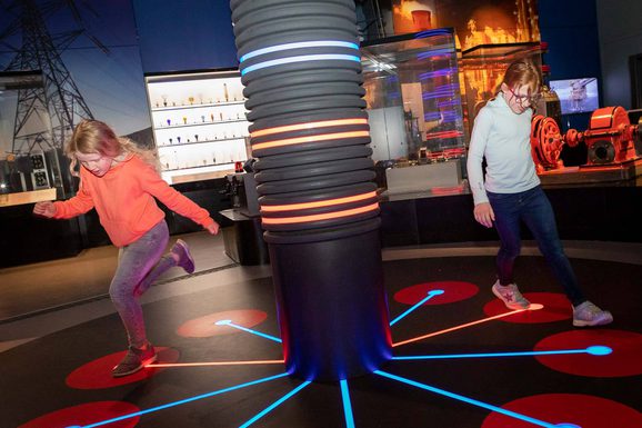Two children run around a lighted pillar in a museum gallery, stepping on different coloured lights on the floor.