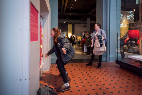 A woman plays an interactive game mounted to a wall in a museum while another woman watches.