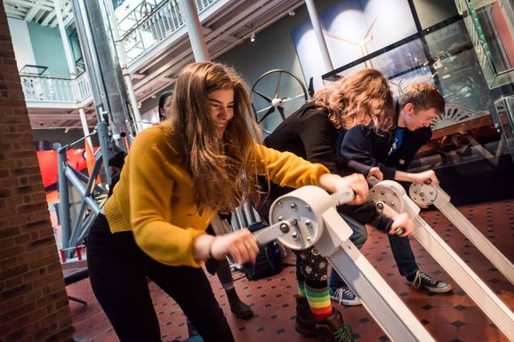 Three adults turning pedals as part of an interactive display in a multi-storey museum gallery.