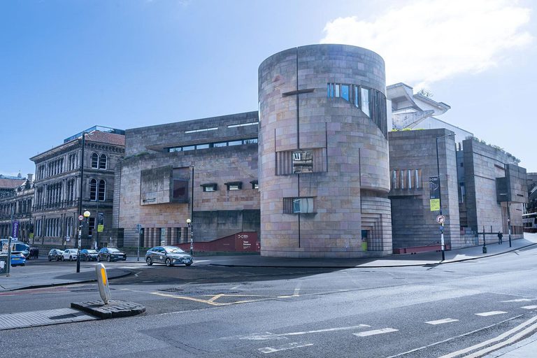The exterior of a large museum building. Cars are parked outside and several people are walking by.