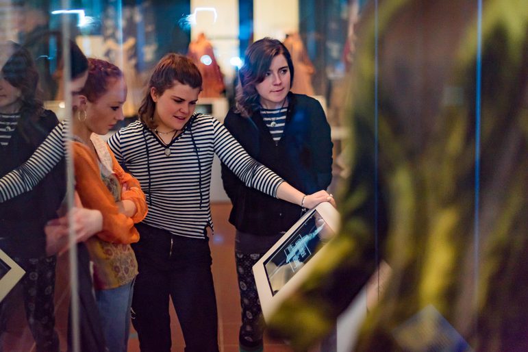 A small group of teenagers look at a screen in a fashion museum.