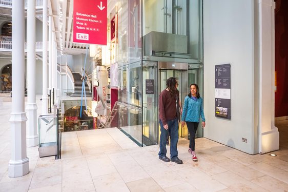 Two visitors walking out of a glass lift in a museum.