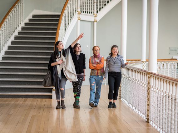 A group of four visitors walk along a balcony in a museum, pointing out different objects on display. There is a set of winding stairs in the background.