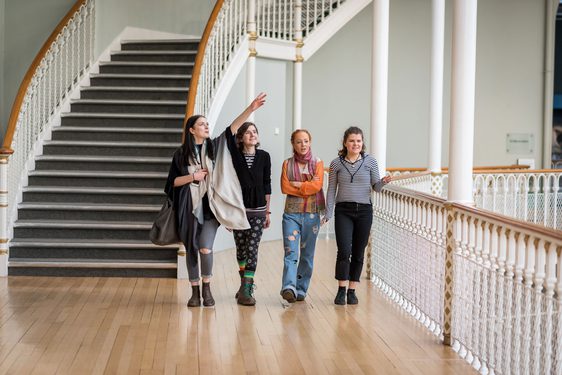 A group of four visitors walk along a balcony in a museum, pointing out different objects on display. There is a set of winding stairs in the background.