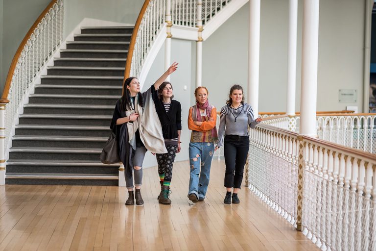 A group of four visitors walk along a balcony in a museum, pointing out different objects on display. There is a set of winding stairs in the background.