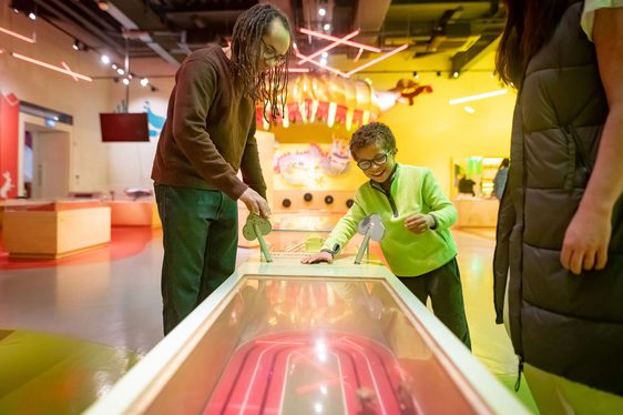 A parent and child interacting with a racing game in a bright and colourful museum gallery.