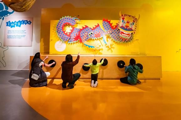 A family of four turning wheels to move a colourful dragon model on a wall above.