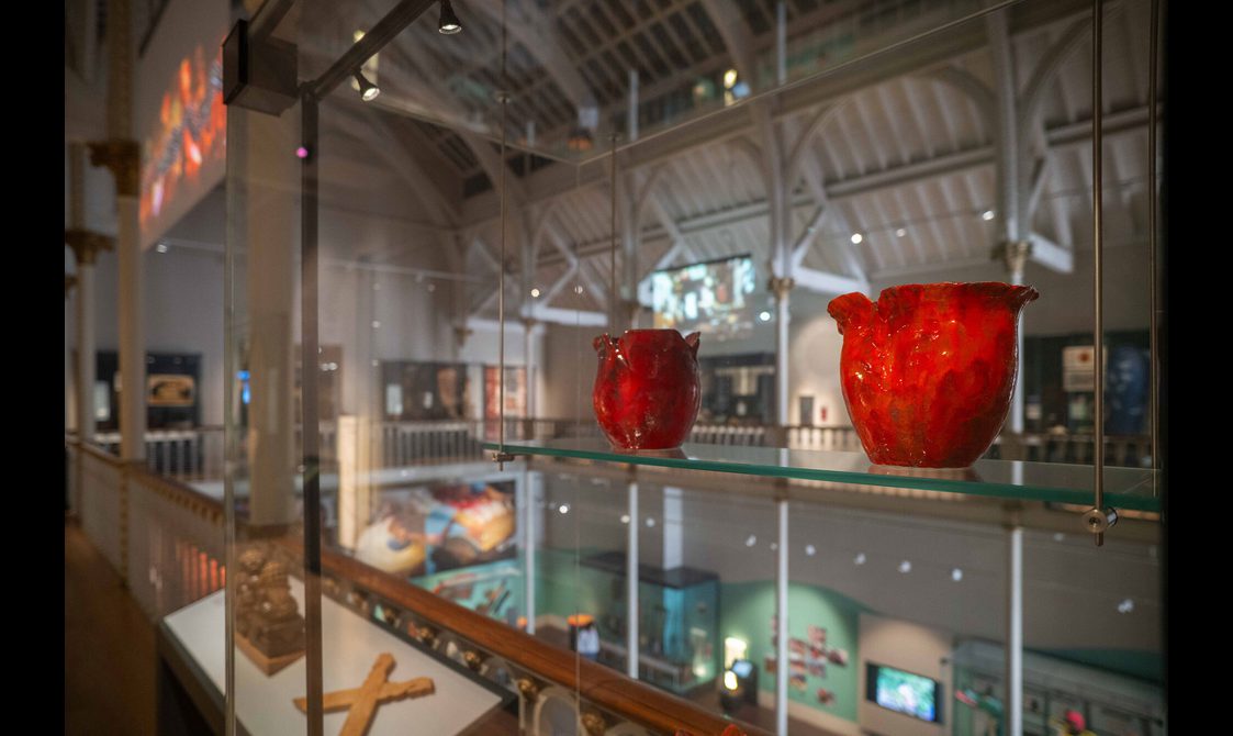 Close up of red ceramic poppy sculptures in a glass case on display in a museum gallery. In the background is a multi-level museum gallery with an arched roof.