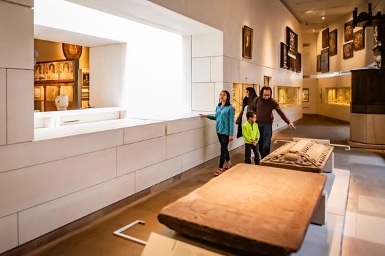 A family walking by two tomb stones in museum gallery. Several portraits are hanging on the walls.