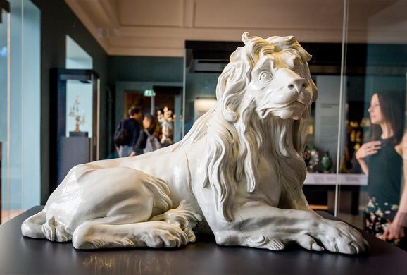 Close up of a porcelain lion figure in a large glass museum case.