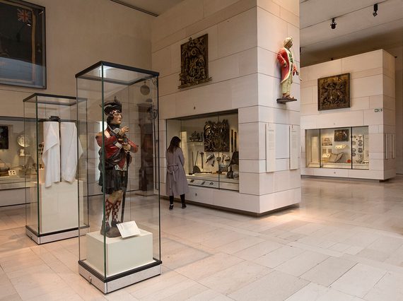 A visitor looking at objects in a glass case in a large museum gallery. There is a sculpture of a soldier in Scottish dress in a cabinet behind the visitor.