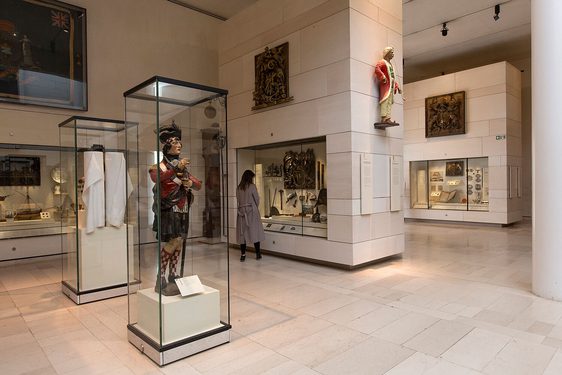 A visitor looking at objects in a glass case in a large museum gallery. There is a sculpture of a soldier in Scottish dress in a cabinet behind the visitor.