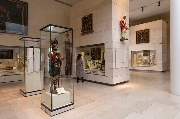 A visitor looking at objects in a glass case in a large museum gallery. There is a sculpture of a soldier in Scottish dress in a cabinet behind the visitor.