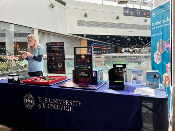 A person stands behind a stall that has different maths and magic activities on it. The stall is in a shopping centre.
