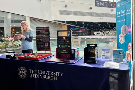 A person stands behind a stall that has different maths and magic activities on it. The stall is in a shopping centre.