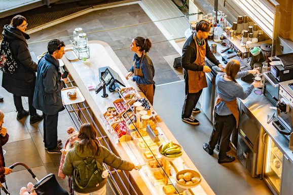 Customers queueing at a cafe till. Baristas are making coffees.
