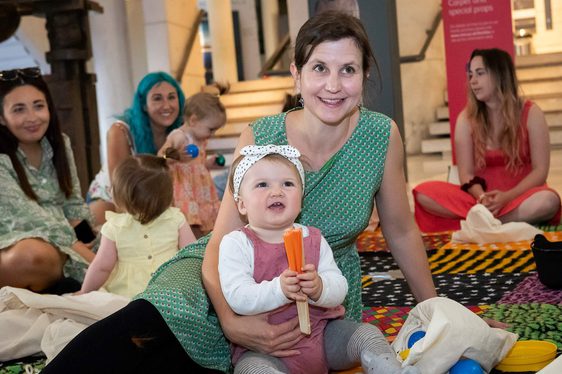 A parent and toddler sitting on a colourful carpet playing with toys. Other parents and toddlers are sitting in the background.