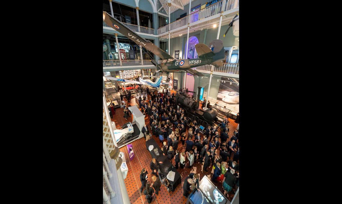 A large group of people at a drinks reception in a multi-level museum gallery. There are different vehicles on display in the gallery and aircrafts suspended from the ceiling.