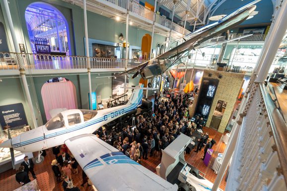 People at a drinks reception in a multi-level museum gallery. There are different aircraft suspended from the ceiling.