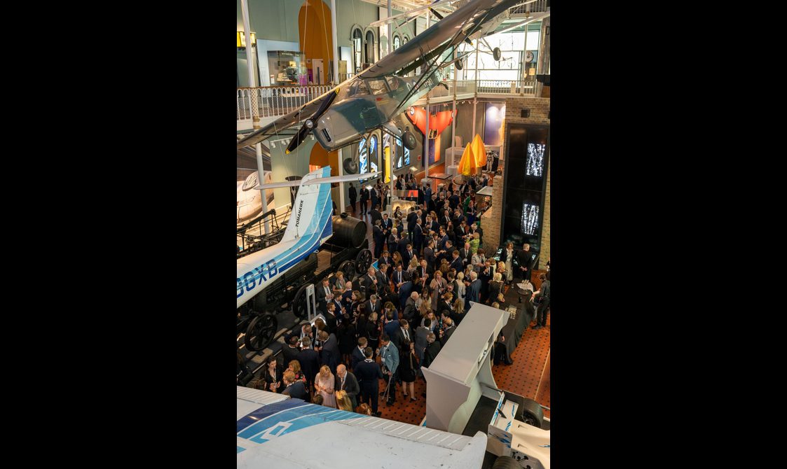 A large group of people at a drinks reception in a multi-level museum gallery. There are different vehicles on display in the gallery and aircrafts suspended from the ceiling.