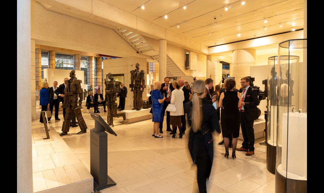 A group of people at a drinks reception in a large museum space with stone floors, high ceilings. There are three brown sculptures of figures in different walking positions.